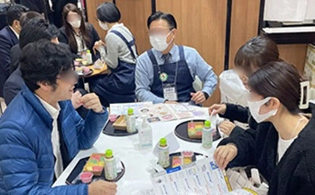 テーブルを囲んで食事をする人々の写真。マスクを着用し、弁当のようなものを食べている。