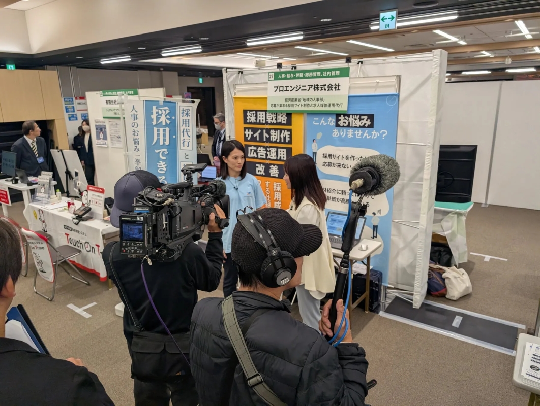 展示会場のような場所で、撮影クルーが「プロエンジニア株式会社」のブースで2人の女性にインタビューを行っている様子です。ブースでは採用戦略やサイト制作、広告運用などの人事関連サービスが紹介されています。