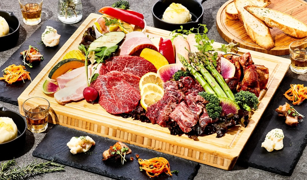 様々なお肉や野菜が盛り付けられた料理の写真です。ステーキ、ソーセージ、野菜、パンなどが木製のプレートに並べられ、周りには小鉢やグラスが配置されています。