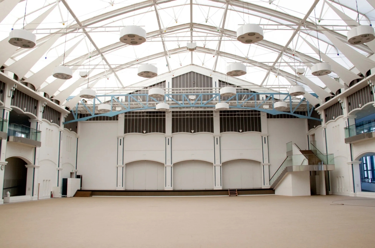 巨大な屋内空間。天井は白く、照明器具が多数設置されている。壁は白く、アーチ型の窓や扉がある。床は明るい茶色で、広いスペースが確保されている。階段やバルコニーも見られる。