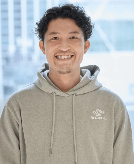 笑顔でカメラを見ている男性のポートレート。男性はグレーのパーカーを着ており、パーカーには「ALL COLORS ARE BEAUTIFUL」というテキストが書かれています。