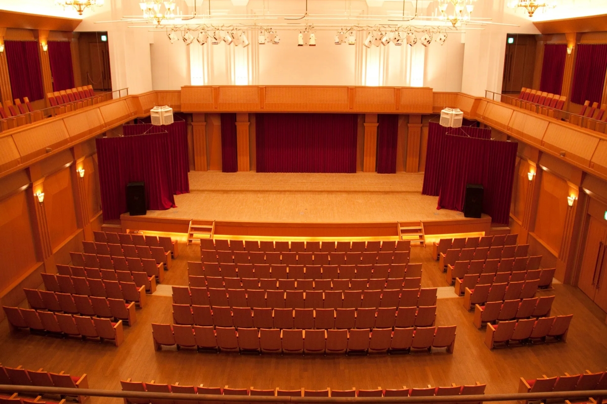 観客席のある劇場です。舞台には赤いカーテンがかけられています。照明が明るく、客席は木製の椅子が並んでいる。