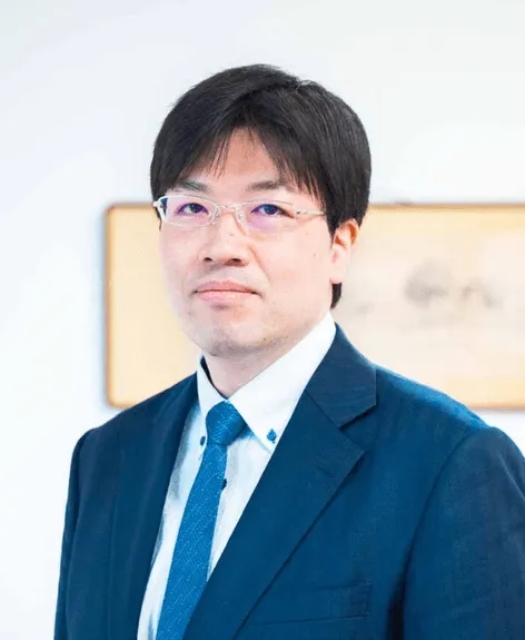 眼鏡をかけた男性のポートレート。彼は濃い色のスーツを着ており、カメラ目線で穏やかな表情をしています。