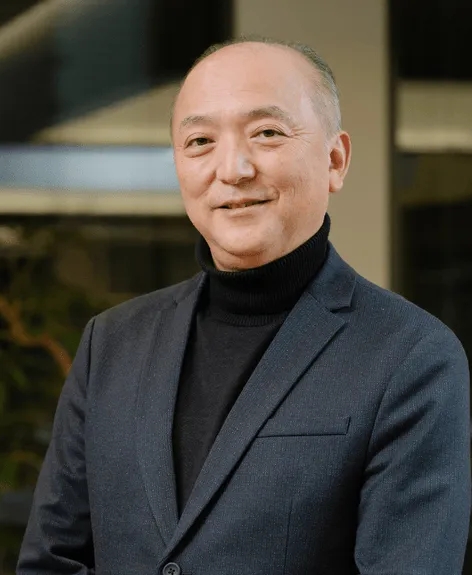 笑顔でカメラを見つめる男性のポートレート。彼は黒いタートルネックセーターとダークブルーのジャケットを着ている。