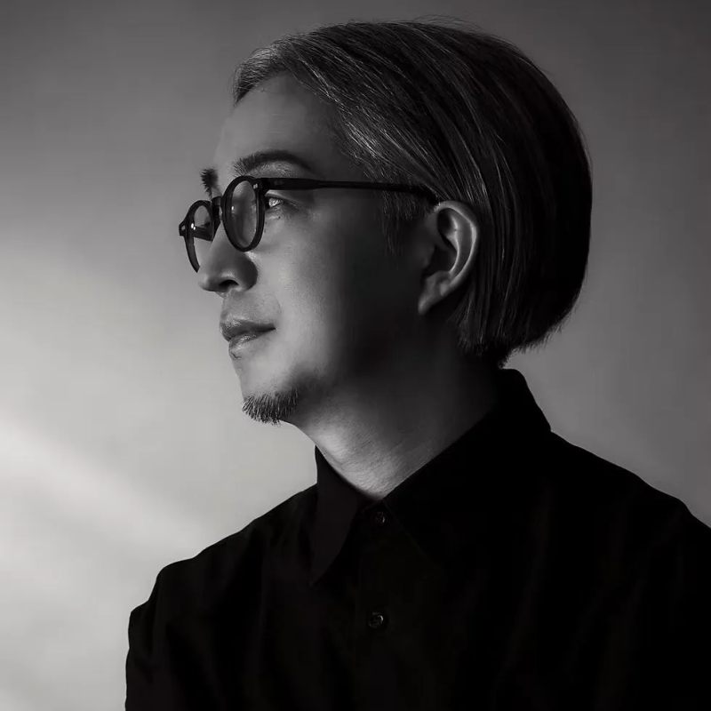 眼鏡をかけた男性のモノクロポートレート。彼は黒いシャツを着ていて、カメラから目をそらしている。