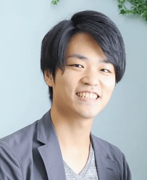 笑顔の男性のポートレートです。彼はジャケットとインナーを着ており、カメラに向かって微笑んでいます。背景は明るい青色で、植物が少し見えます。