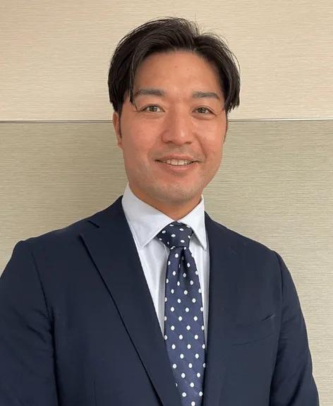 ネイビーのスーツを着た笑顔の男性のポートレートです。白いシャツとドット柄のネクタイをしています。背景は明るい色の木目調の壁です。