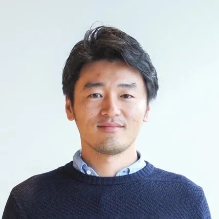 正面を向いたアジア人男性のポートレート。彼は濃い色のセーターと、その下に水色のシャツを着ている。背景は白。