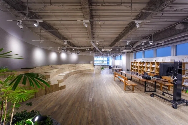 モダンなデザインのオフィスラウンジ。木製の床、階段状の座席、植物、本棚、大型モニターが特徴です。