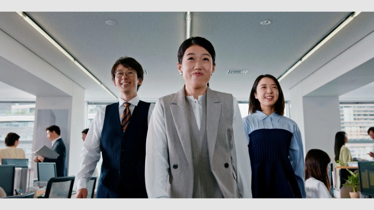 オフィスで笑顔で歩く3人のビジネスパーソン。他の従業員も写っている。