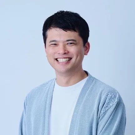 笑顔の男性のポートレートです。彼は白いTシャツと水色のカーディガンを着ています。背景は白です。