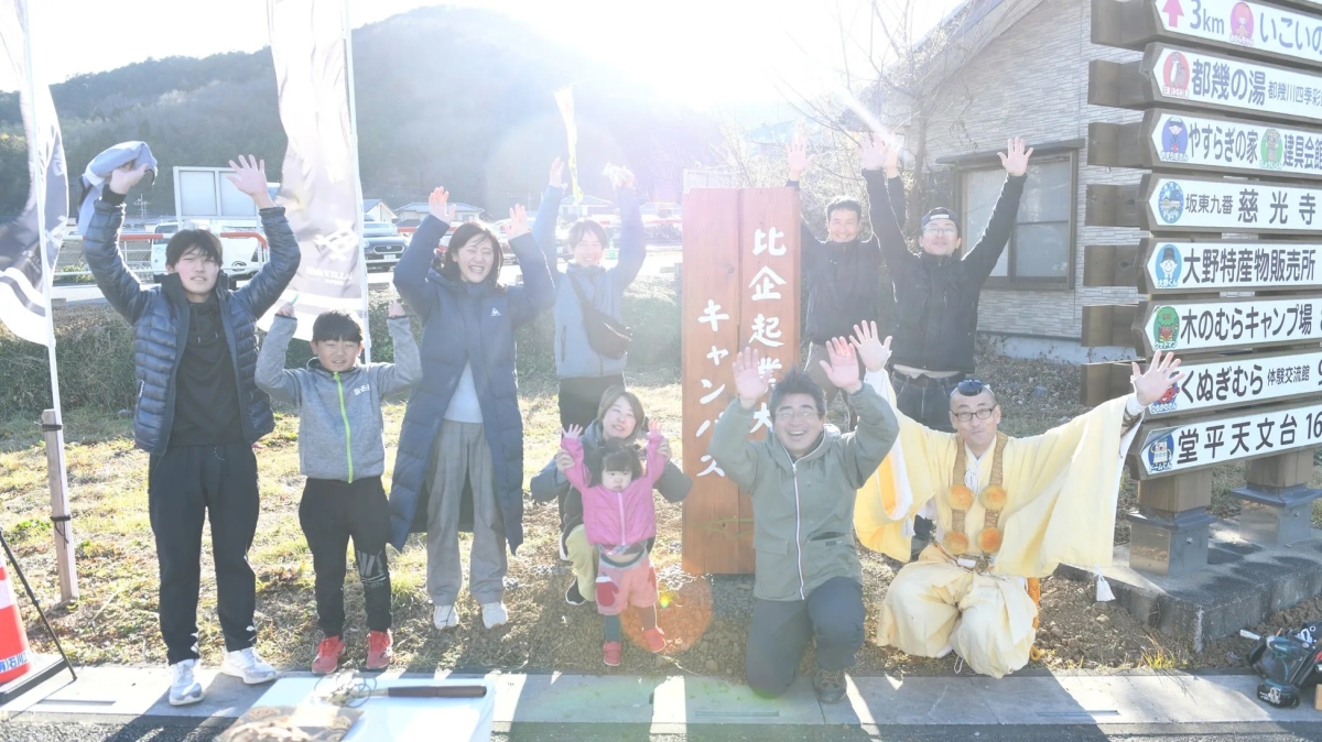 多くの人が手を上げて立っている写真。看板の前に立っており、比企起キャンバスと書かれています。看板の横には、さまざまな施設への距離を示す看板があります。