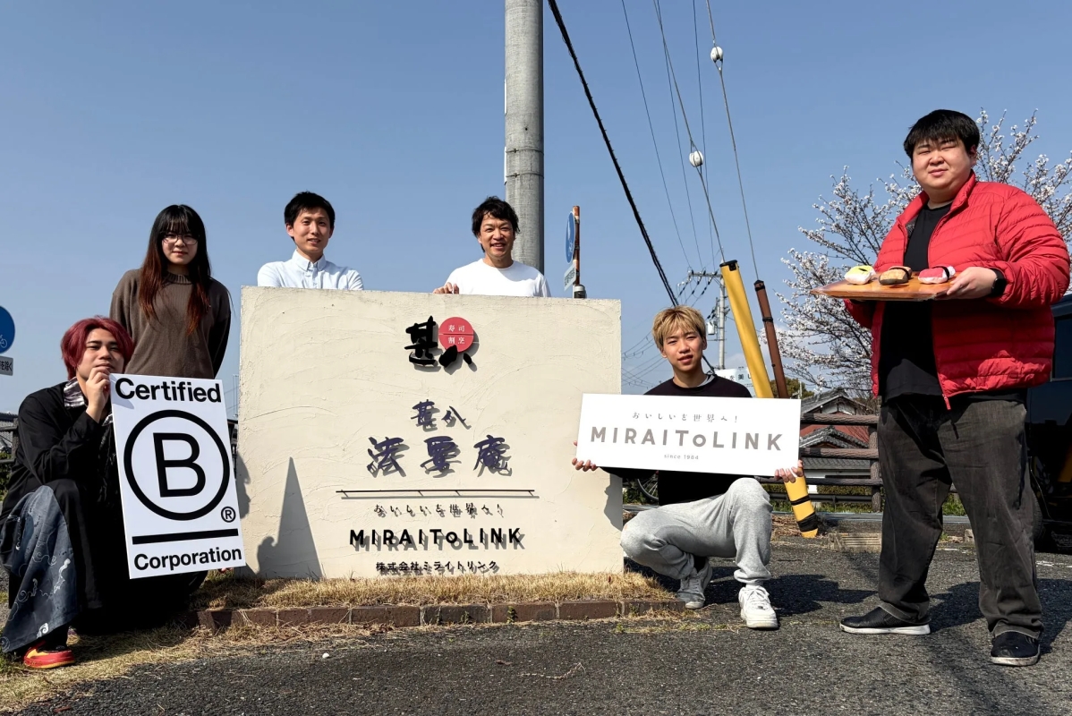 看板の前に立つ人々。看板には「MIRAITOLINK」と「港憂慮」の文字が見える。手前には「Certified B Corporation」のロゴを持つ人がいる。男性が寿司を乗せたお盆を持っている。