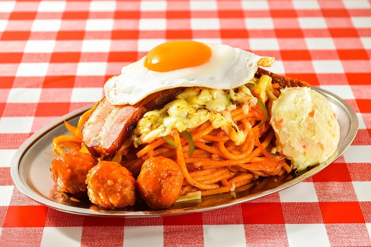 パスタの上に目玉焼き、ベーコン、チーズが乗った料理の写真。ミートボールとポテトサラダも添えられている。